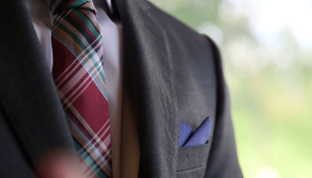 Shoulder of a well dressed man in a black suit with a plaid tie and navy blue pocket square.